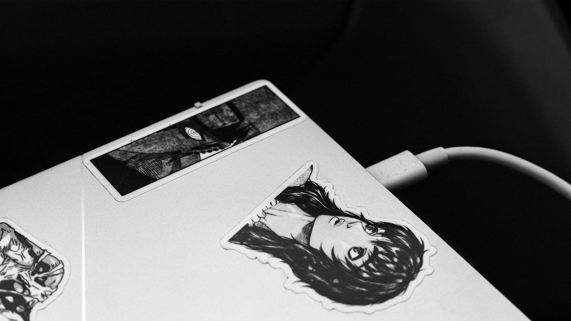 Laptop z ładowarką, ozdobiony czarno-białymi naklejkami z anime
